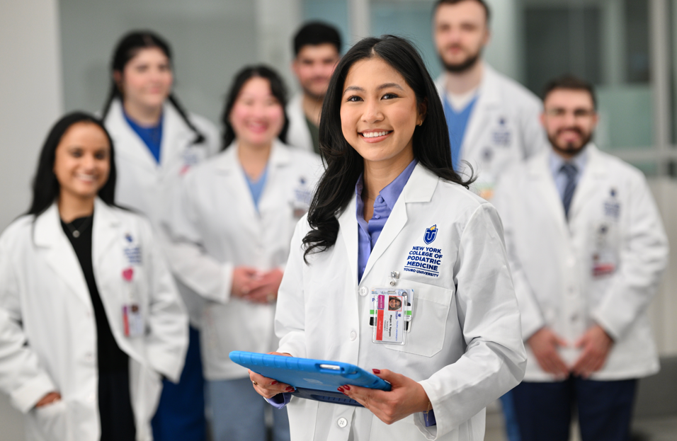 A group of Touro New York College of Podiatric Medicine students wearing white coats, with one female student in front holding a tablet.